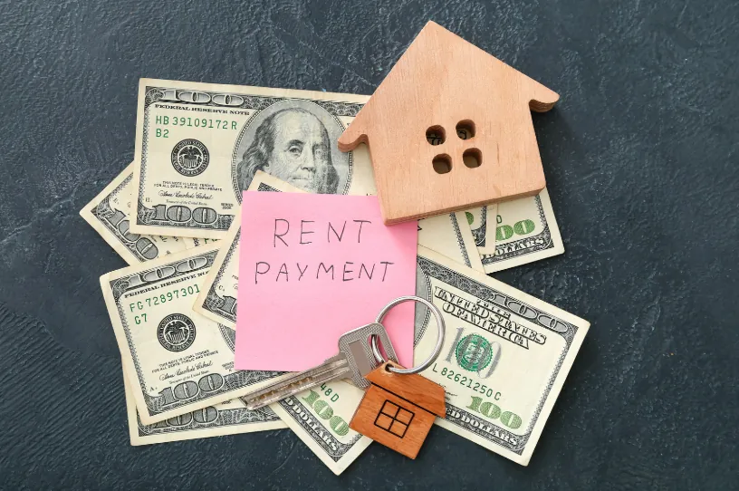 A small wooden house model, cash, and keys on a dark surface with a note that says "Rent Payment," emphasizing financial planning for how much rent can i afford.