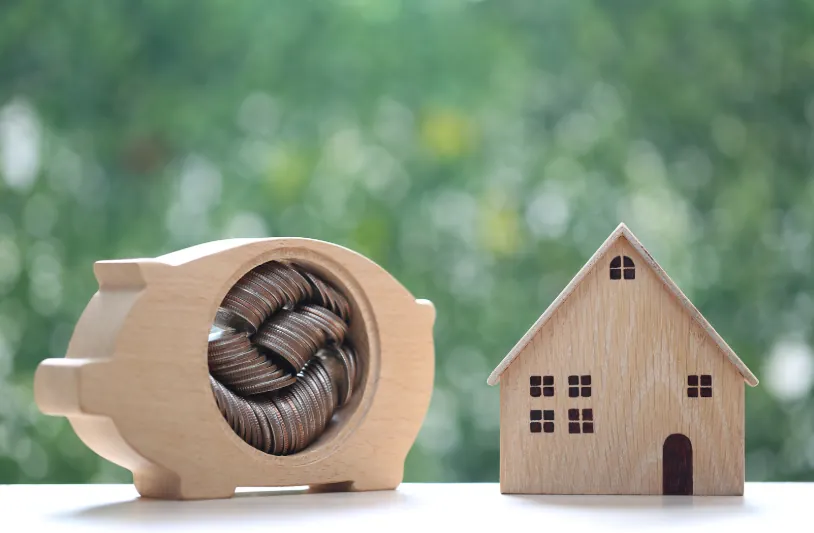 A wooden piggy bank filled with coins next to a small house model, representing savings for home expenses, rent, and the pet fee.